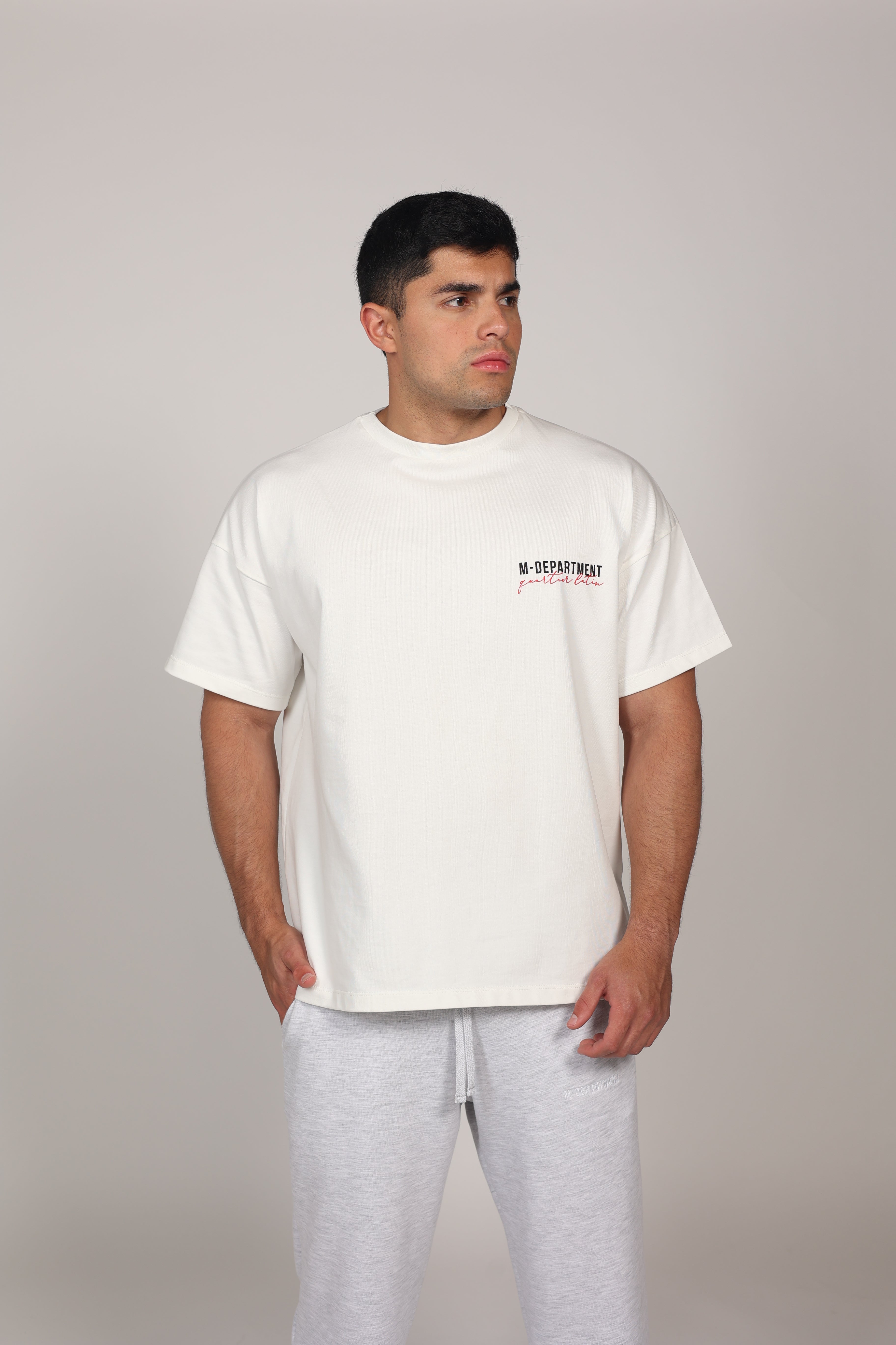 Modelo masculino posando con una camiseta oversize blanca de algodón 100%. La prenda presenta hombros caídos, mangas amplias y el texto 'M-DEPARTMENT' en el lado izquierdo del pecho en letras negras y rojas. El modelo complementa el look con un pantalón deportivo gris claro sobre un fondo neutro, resaltando una estética urbana y deportiva."