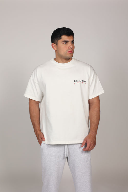 Modelo masculino posando con una camiseta oversize blanca de algodón 100%. La prenda presenta hombros caídos, mangas amplias y el texto 'M-DEPARTMENT' en el lado izquierdo del pecho en letras negras y rojas. El modelo complementa el look con un pantalón deportivo gris claro sobre un fondo neutro, resaltando una estética urbana y deportiva."