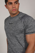 Hombre atlético de perfil vistiendo una camiseta de corte raglán en color gris jaspeado. La prenda destaca por sus costuras diagonales desde el cuello hasta la sisa, resaltando la musculatura de los hombros y el pecho. En el centro del pecho se aprecia un pequeño branding numérico sutil sobre un fondo gris claro neutro.