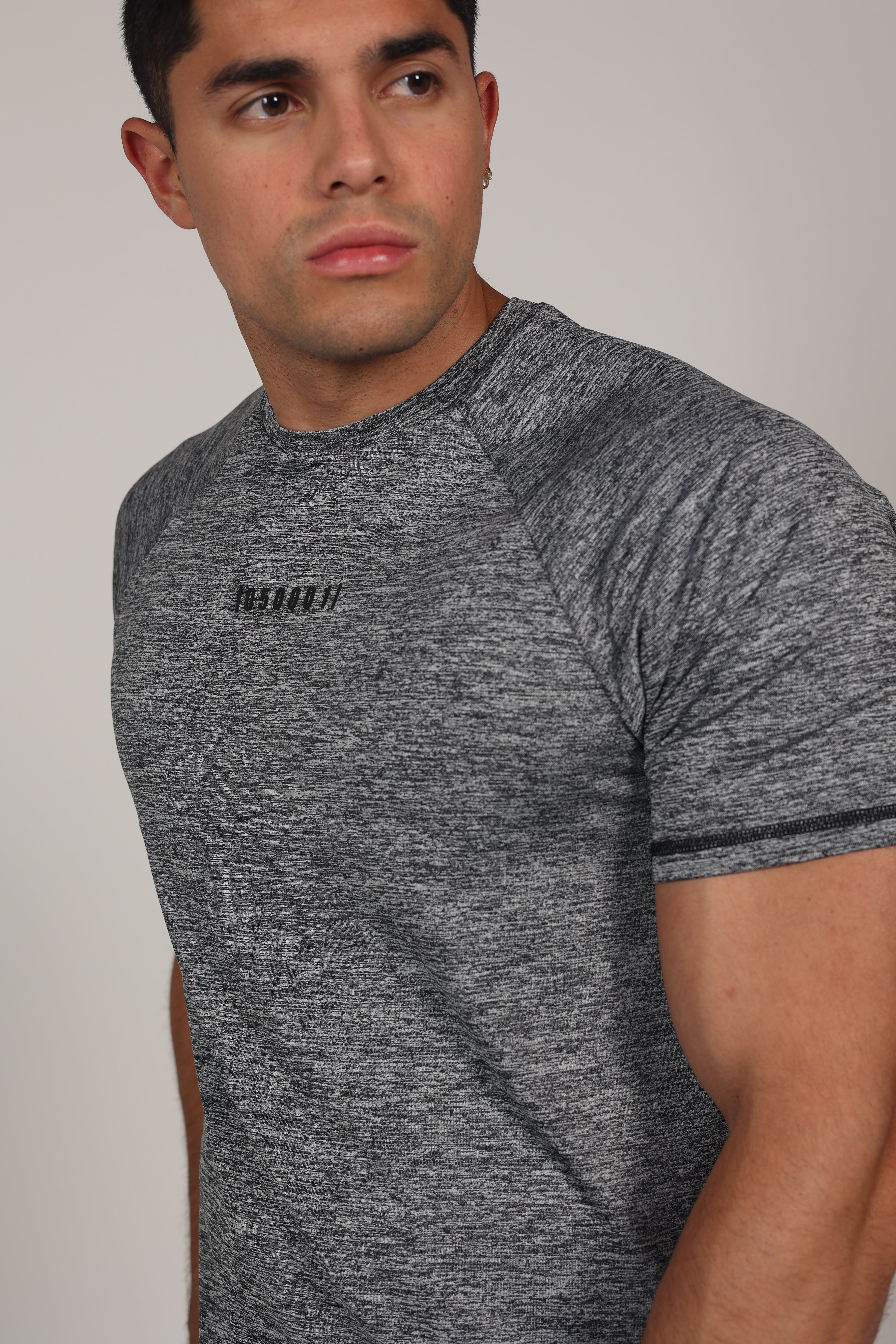 Hombre atlético de perfil vistiendo una camiseta de corte raglán en color gris jaspeado. La prenda destaca por sus costuras diagonales desde el cuello hasta la sisa, resaltando la musculatura de los hombros y el pecho. En el centro del pecho se aprecia un pequeño branding numérico sutil sobre un fondo gris claro neutro.