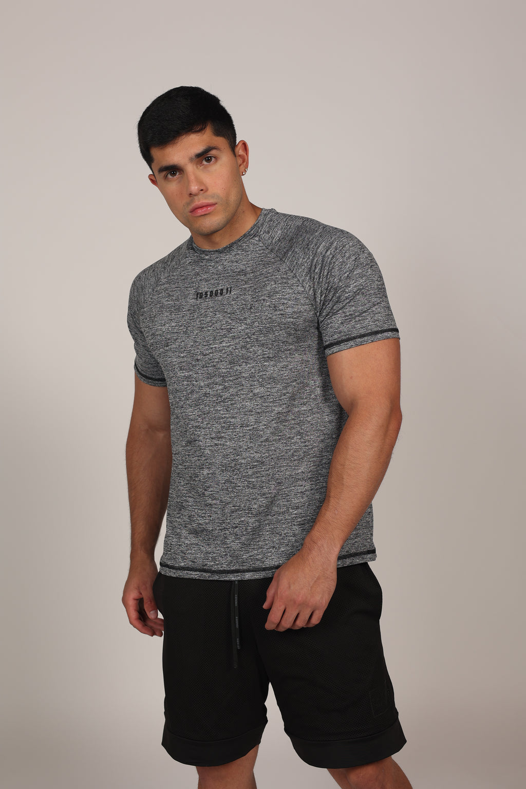 Hombre atlético de perfil vistiendo una camiseta de corte raglán en color gris jaspeado. La prenda destaca por sus costuras diagonales desde el cuello hasta la sisa, resaltando la musculatura de los hombros y el pecho. En el centro del pecho se aprecia un pequeño branding numérico sutil sobre un fondo gris claro neutro.