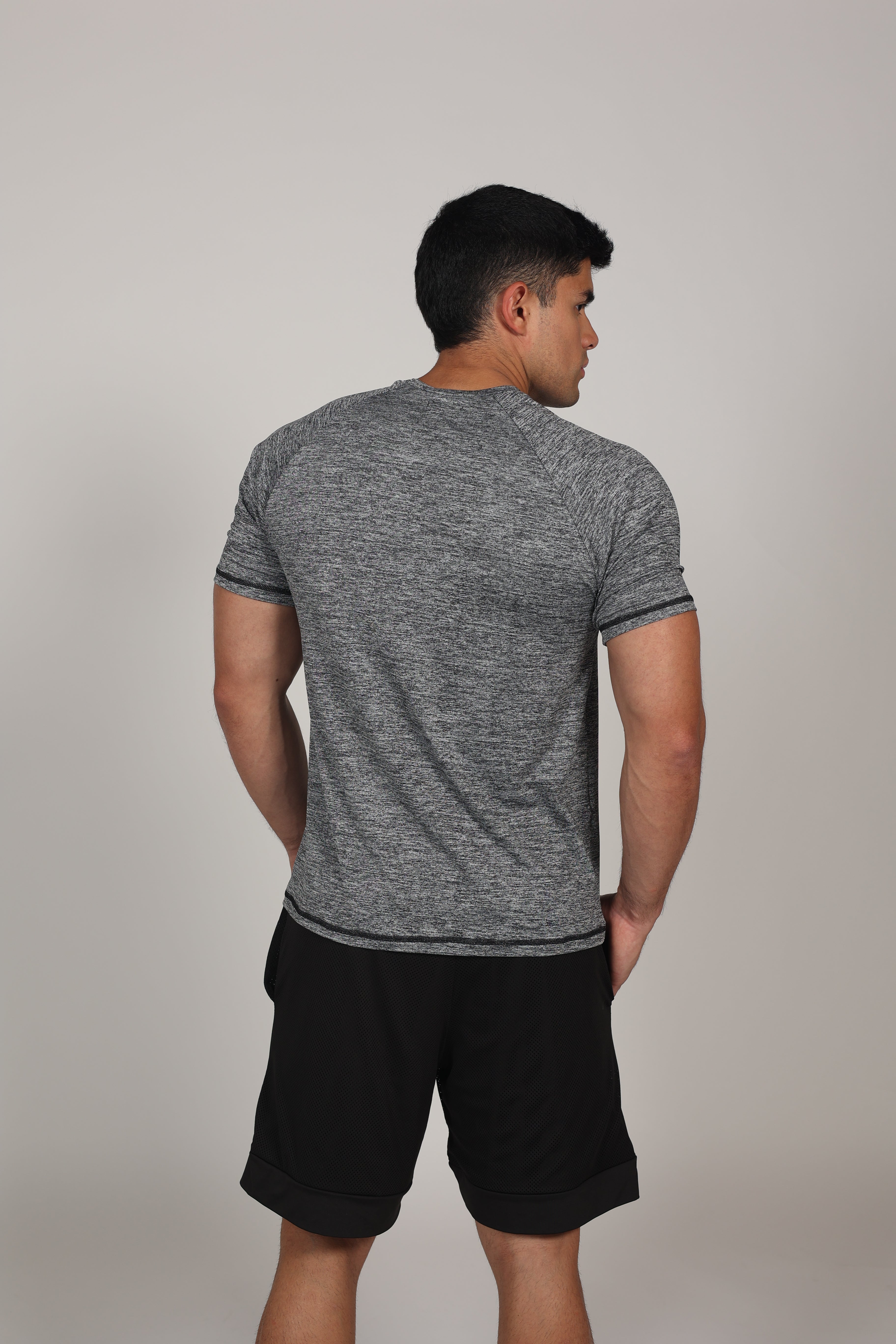 Hombre atlético de perfil vistiendo una camiseta de corte raglán en color gris jaspeado. La prenda destaca por sus costuras diagonales desde el cuello hasta la sisa, resaltando la musculatura de los hombros y el pecho. En el centro del pecho se aprecia un pequeño branding numérico sutil sobre un fondo gris claro neutro.