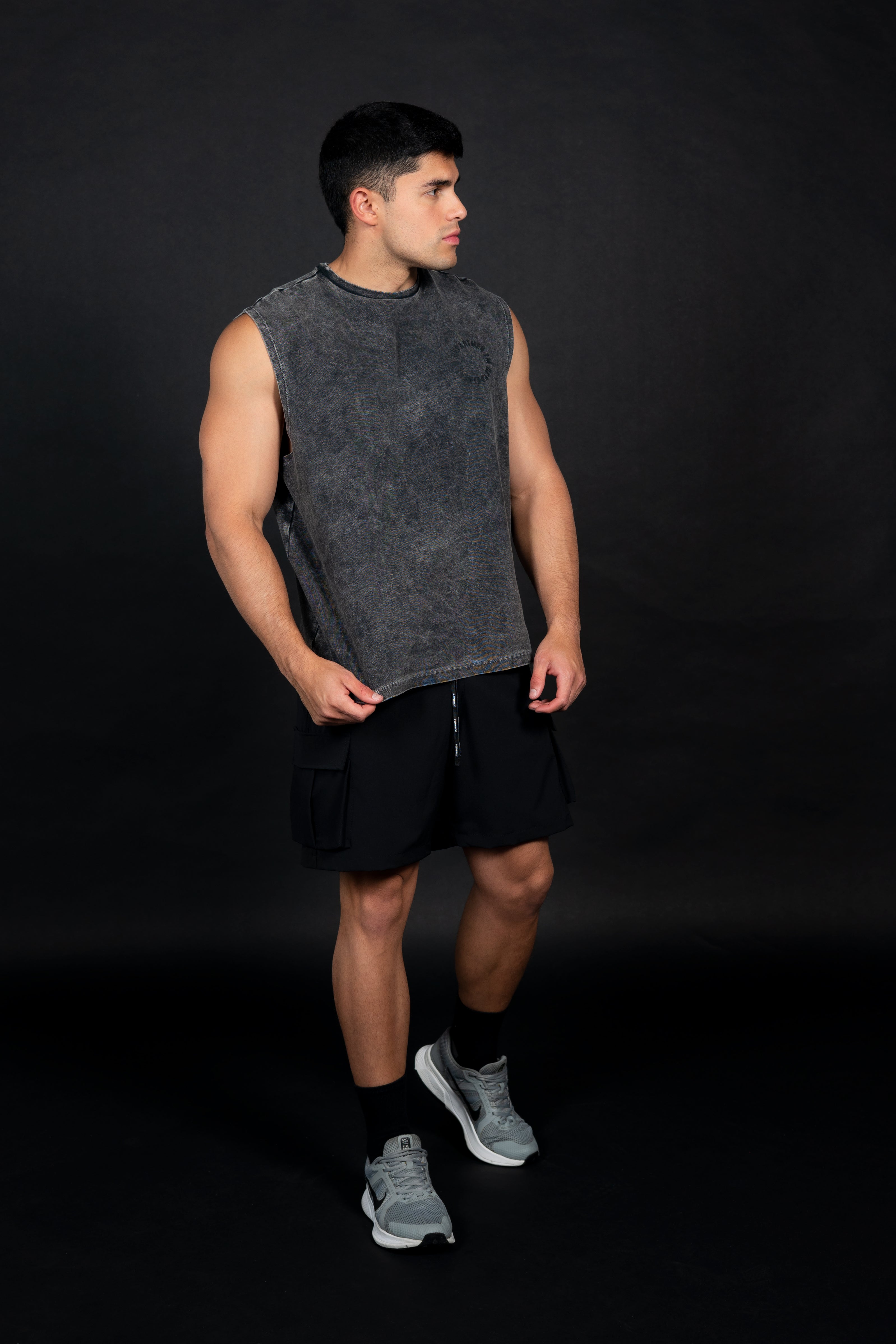 Modelo masculino atlético de perfil usando una camiseta sin mangas (muscle tee) de color gris oscuro con efecto de lavado vintage y un pequeño logo circular en el pecho. Viste pantalones cortos cargo negros y zapatillas deportivas grises, posando sobre un fondo negro liso que resalta la textura de la prenda.