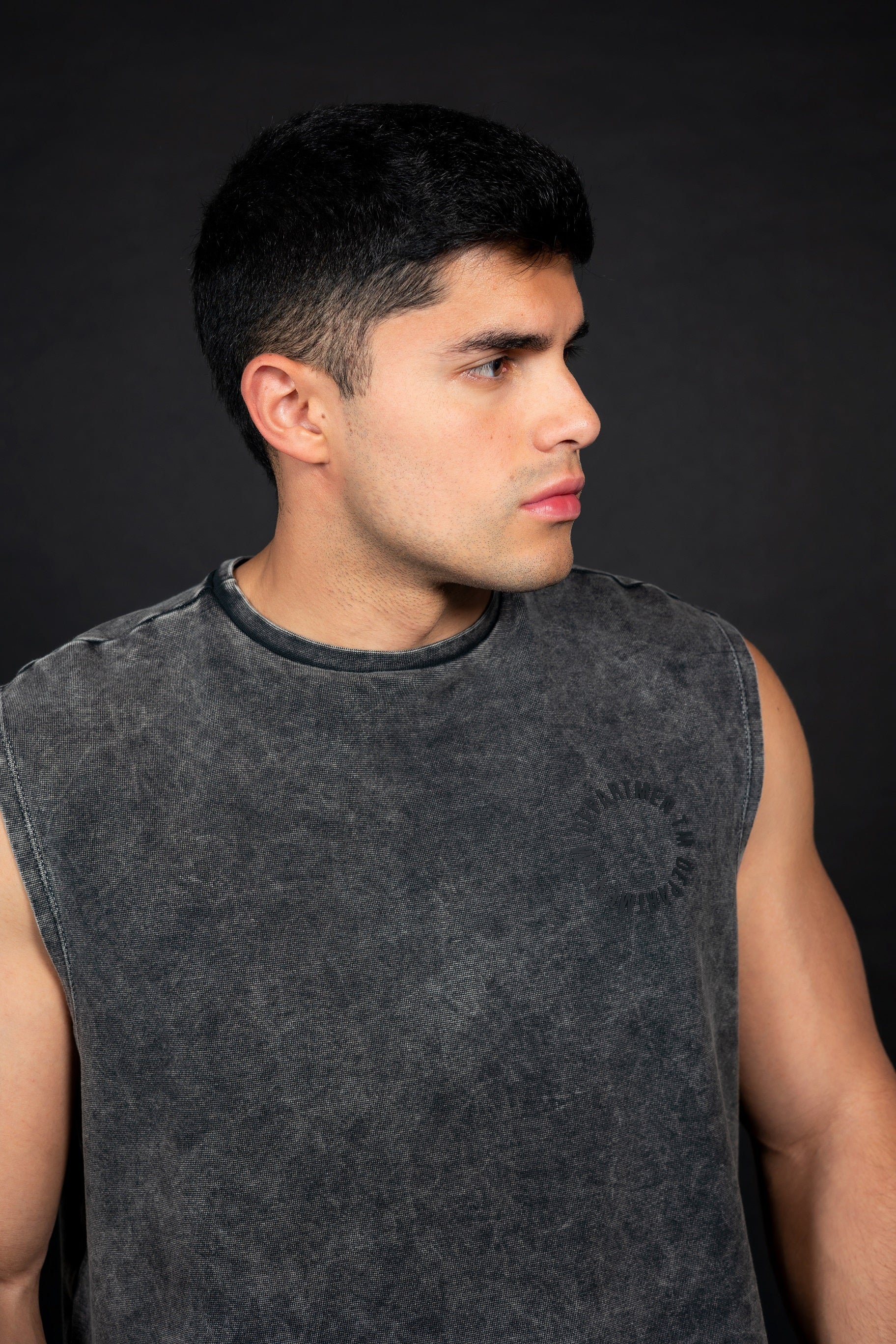 Modelo masculino atlético de perfil usando una camiseta sin mangas (muscle tee) de color gris oscuro con efecto de lavado vintage y un pequeño logo circular en el pecho. Viste pantalones cortos cargo negros y zapatillas deportivas grises, posando sobre un fondo negro liso que resalta la textura de la prenda.