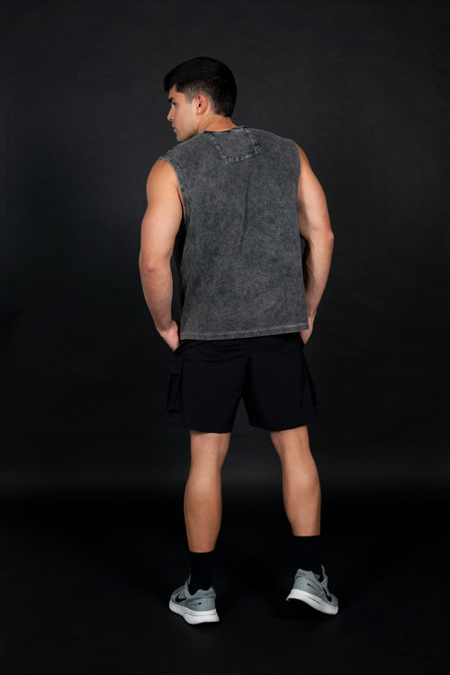 Modelo masculino atlético de perfil usando una camiseta sin mangas (muscle tee) de color gris oscuro con efecto de lavado vintage y un pequeño logo circular en el pecho. Viste pantalones cortos cargo negros y zapatillas deportivas grises, posando sobre un fondo negro liso que resalta la textura de la prenda.