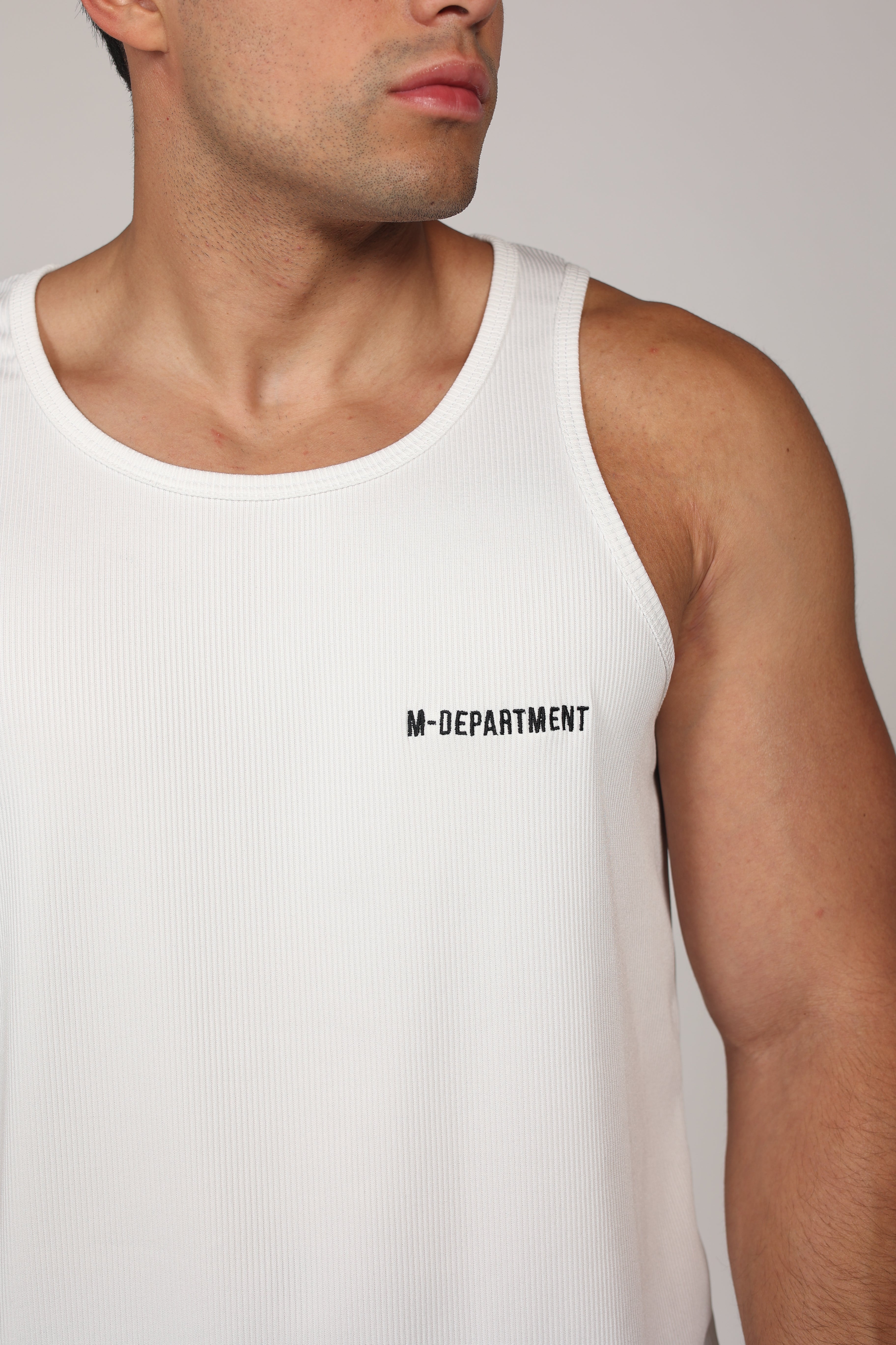 Primer plano de un hombre atlético vistiendo una camiseta sin mangas blanca de tela acanalada. La prenda presenta el texto 'M-DEPARTMENT' en letras negras pequeñas en el lado izquierdo del pecho. El fondo es de un tono gris neutro claro, destacando la textura del tejido y el ajuste atlético de la prenda.