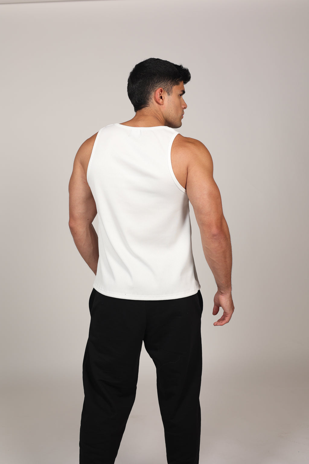 Primer plano de un hombre atlético vistiendo una camiseta sin mangas blanca de tela acanalada. La prenda presenta el texto 'M-DEPARTMENT' en letras negras pequeñas en el lado izquierdo del pecho. El fondo es de un tono gris neutro claro, destacando la textura del tejido y el ajuste atlético de la prenda.