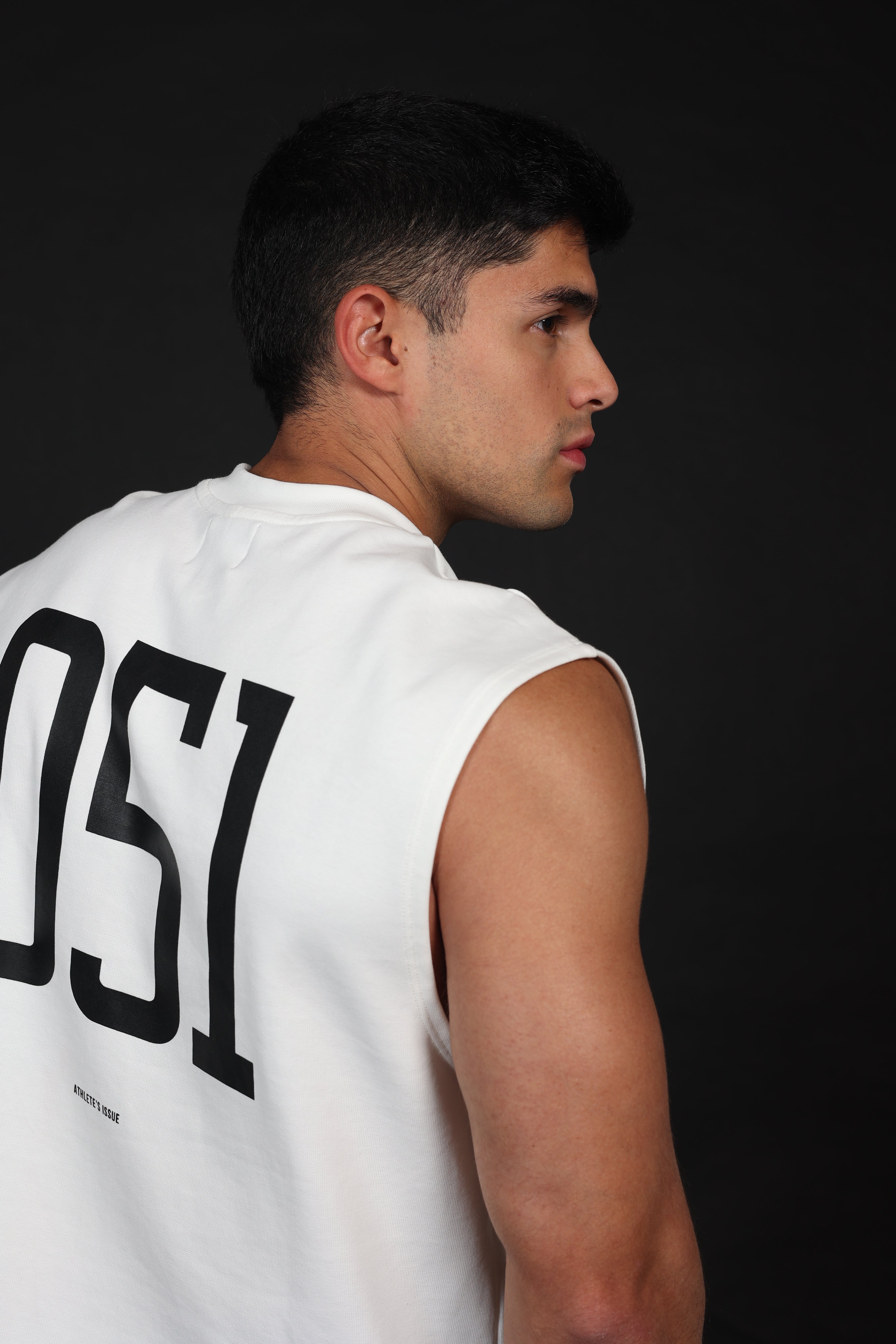 Primer plano de una camiseta sin mangas para hombre en color blanco, confeccionada en algodón elástico con textura acanalada vertical. En el lado izquierdo del pecho se lee el texto 'M-DEPARTMENT' en letras negras minimalistas. La prenda muestra un cuello redondo reforzado y un ajuste atlético que resalta los hombros y el pecho.