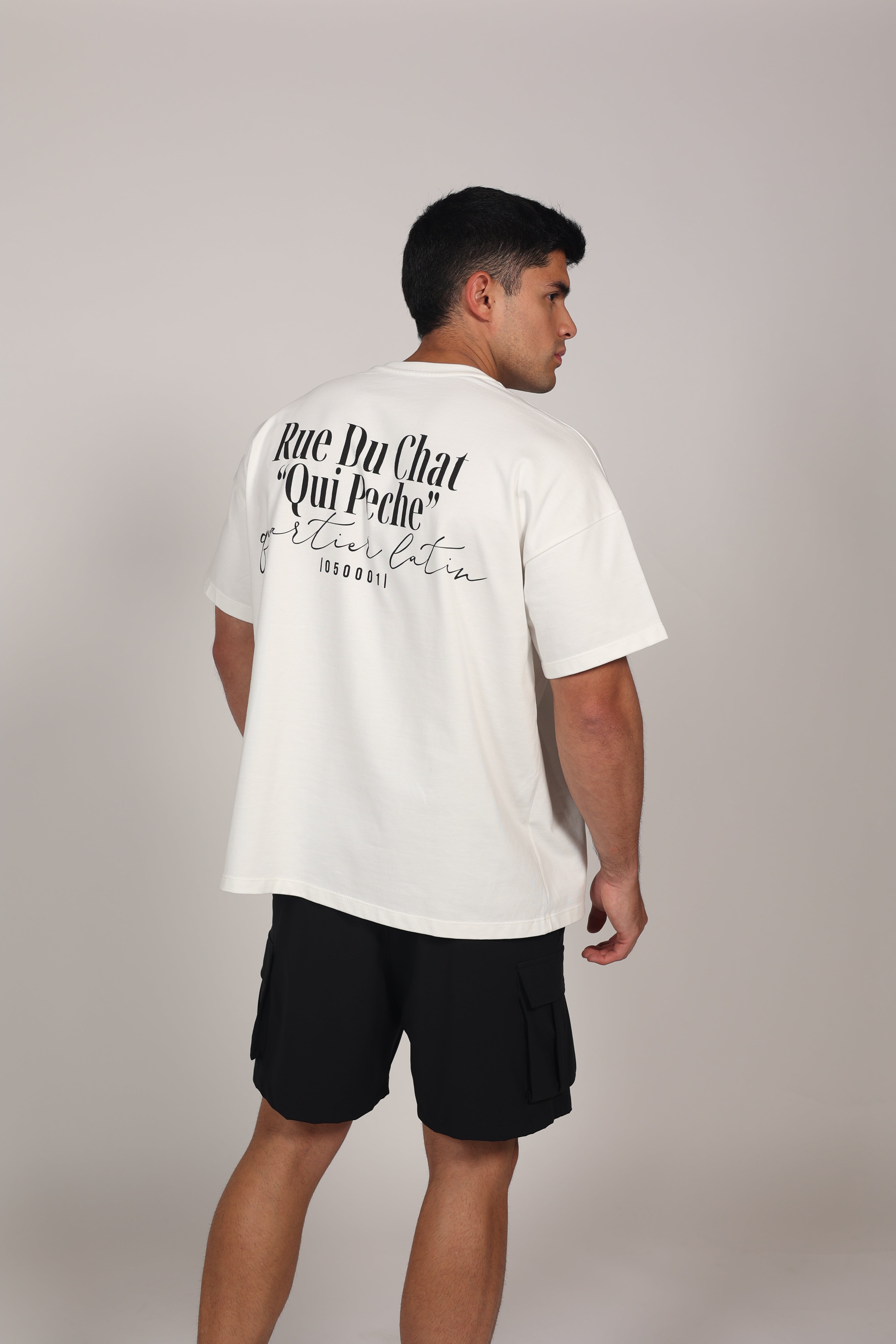Modelo masculino posando con una camiseta oversize blanca de algodón 100%. La prenda presenta hombros caídos, mangas amplias y el texto 'M-DEPARTMENT' en el lado izquierdo del pecho en letras negras y rojas. El modelo complementa el look con un pantalón deportivo gris claro sobre un fondo neutro, resaltando una estética urbana y deportiva."