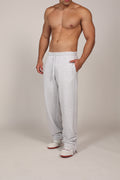 Hombre atlético vistiendo un pantalón deportivo jogger en color gris claro jaspeado. La prenda cuenta con una cintura elástica fruncida con cordón de ajuste. El corte es relajado y se muestra combinado con unos sneakers blancos con detalles rojos, destacando su caída moderna sobre el calzado en un fondo gris neutro o negro