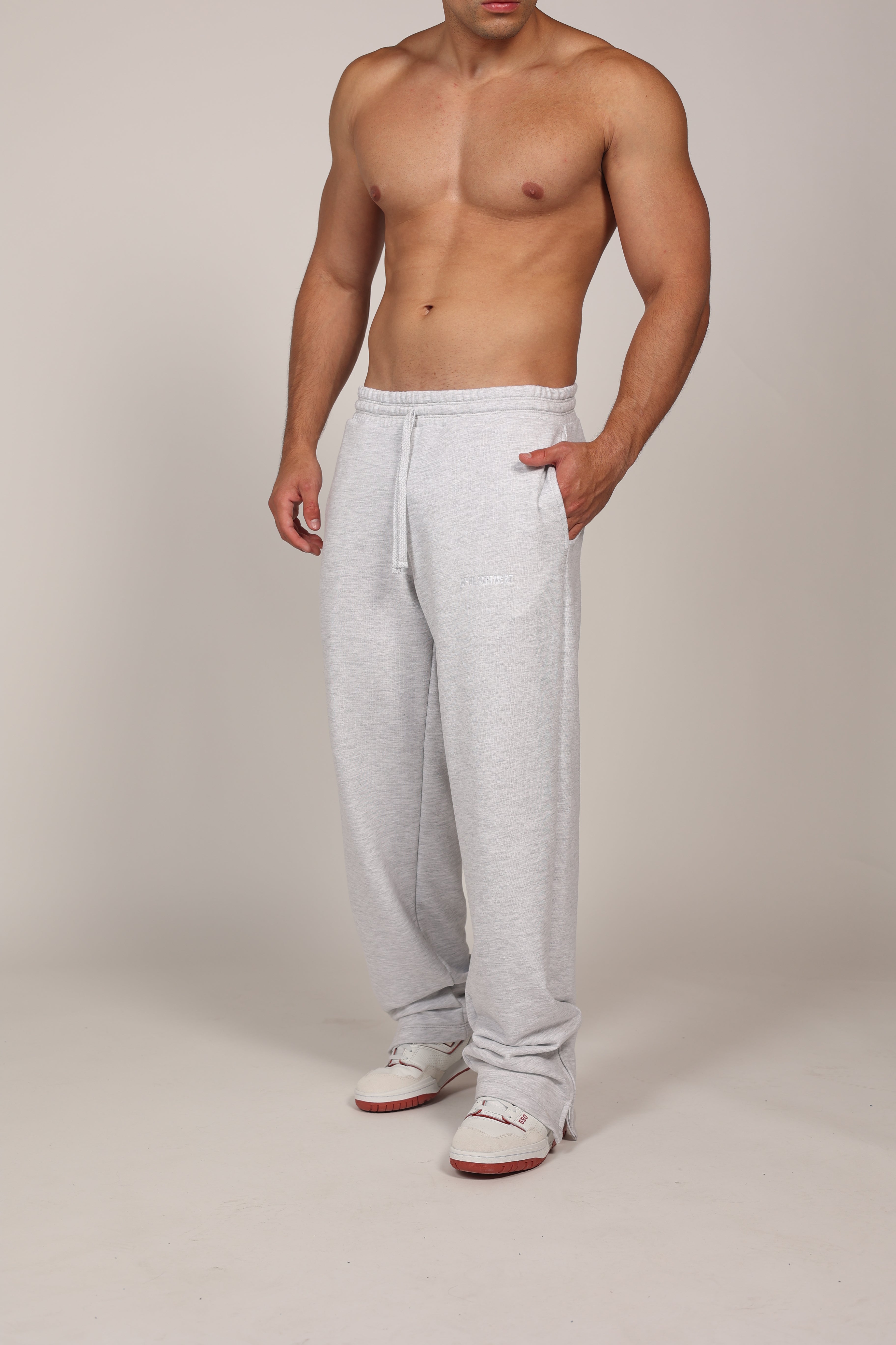 Hombre atlético vistiendo un pantalón deportivo jogger en color gris claro jaspeado. La prenda cuenta con una cintura elástica fruncida con cordón de ajuste. El corte es relajado y se muestra combinado con unos sneakers blancos con detalles rojos, destacando su caída moderna sobre el calzado en un fondo gris neutro o negro