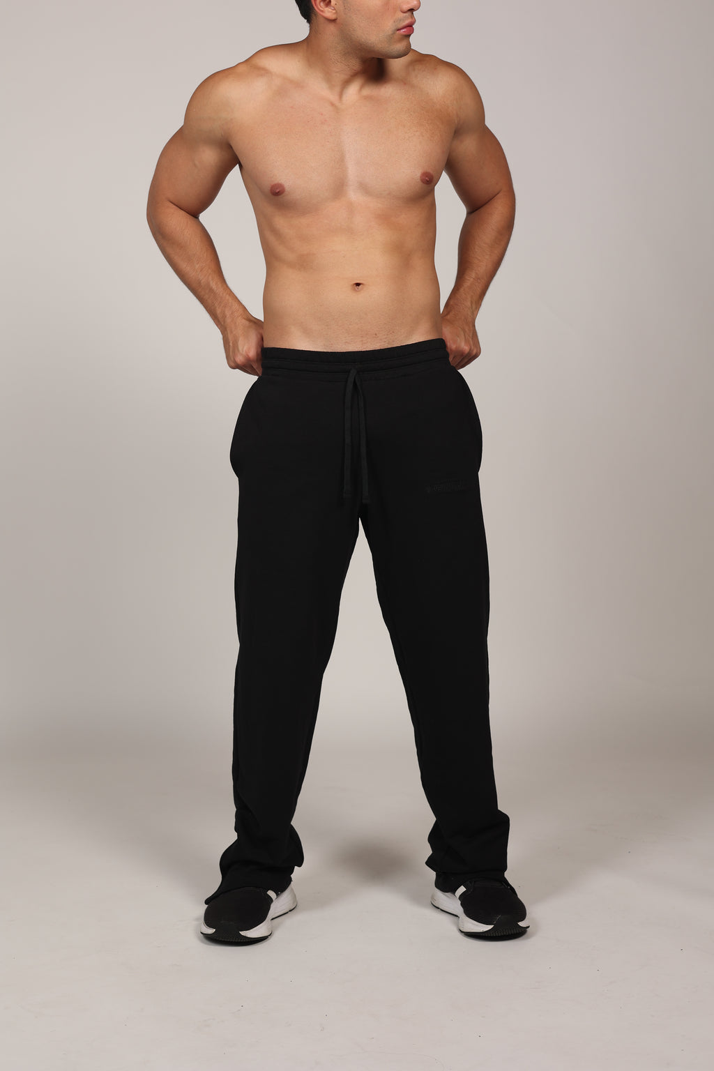 Hombre atlético vistiendo un pantalón deportivo jogger en color gris claro jaspeado. La prenda cuenta con una cintura elástica fruncida con cordón de ajuste. El corte es relajado y se muestra combinado con unos sneakers blancos con detalles rojos, destacando su caída moderna sobre el calzado en un fondo gris neutro o negro