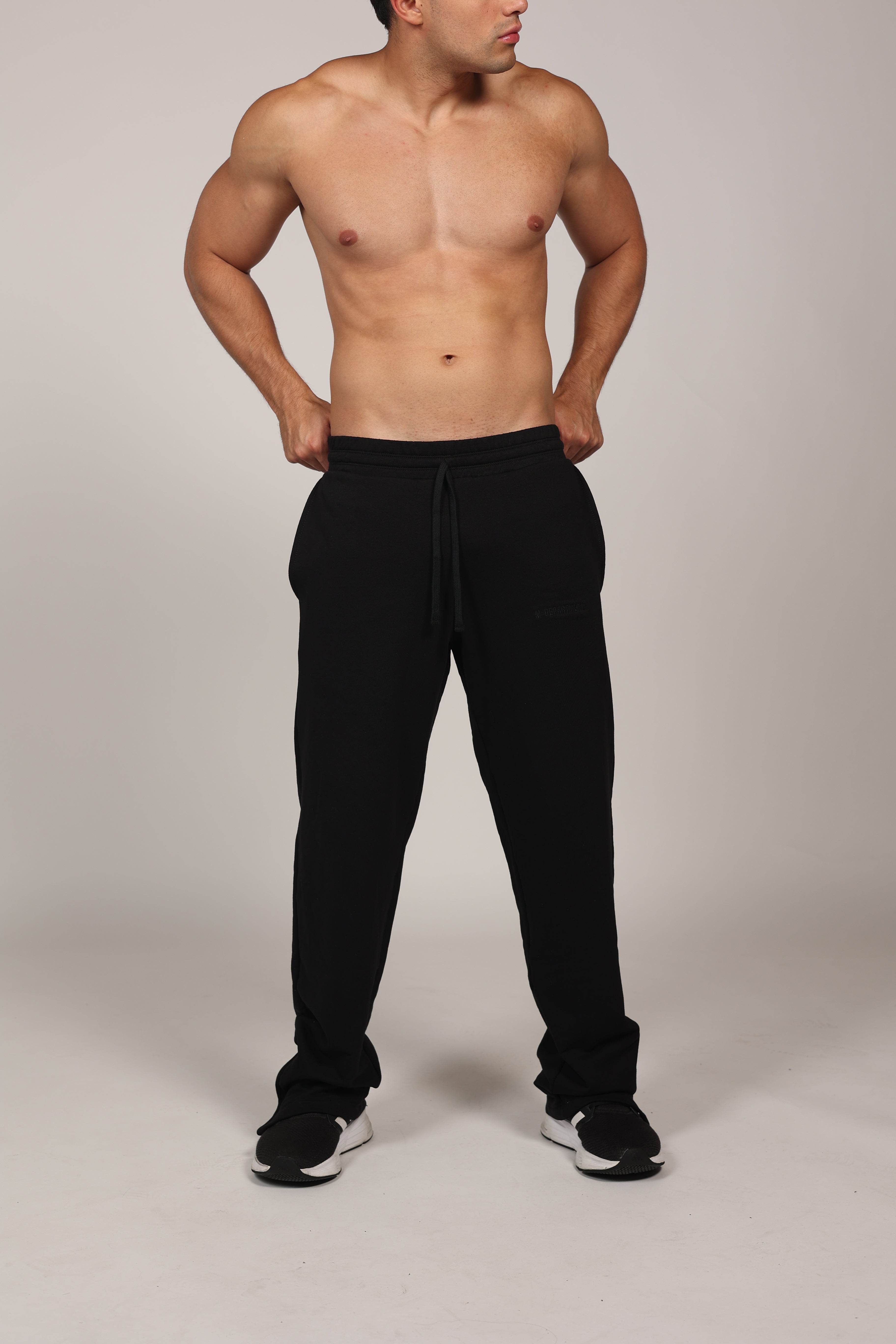 Hombre atlético vistiendo un pantalón deportivo jogger en color gris claro jaspeado. La prenda cuenta con una cintura elástica fruncida con cordón de ajuste. El corte es relajado y se muestra combinado con unos sneakers blancos con detalles rojos, destacando su caída moderna sobre el calzado en un fondo gris neutro o negro