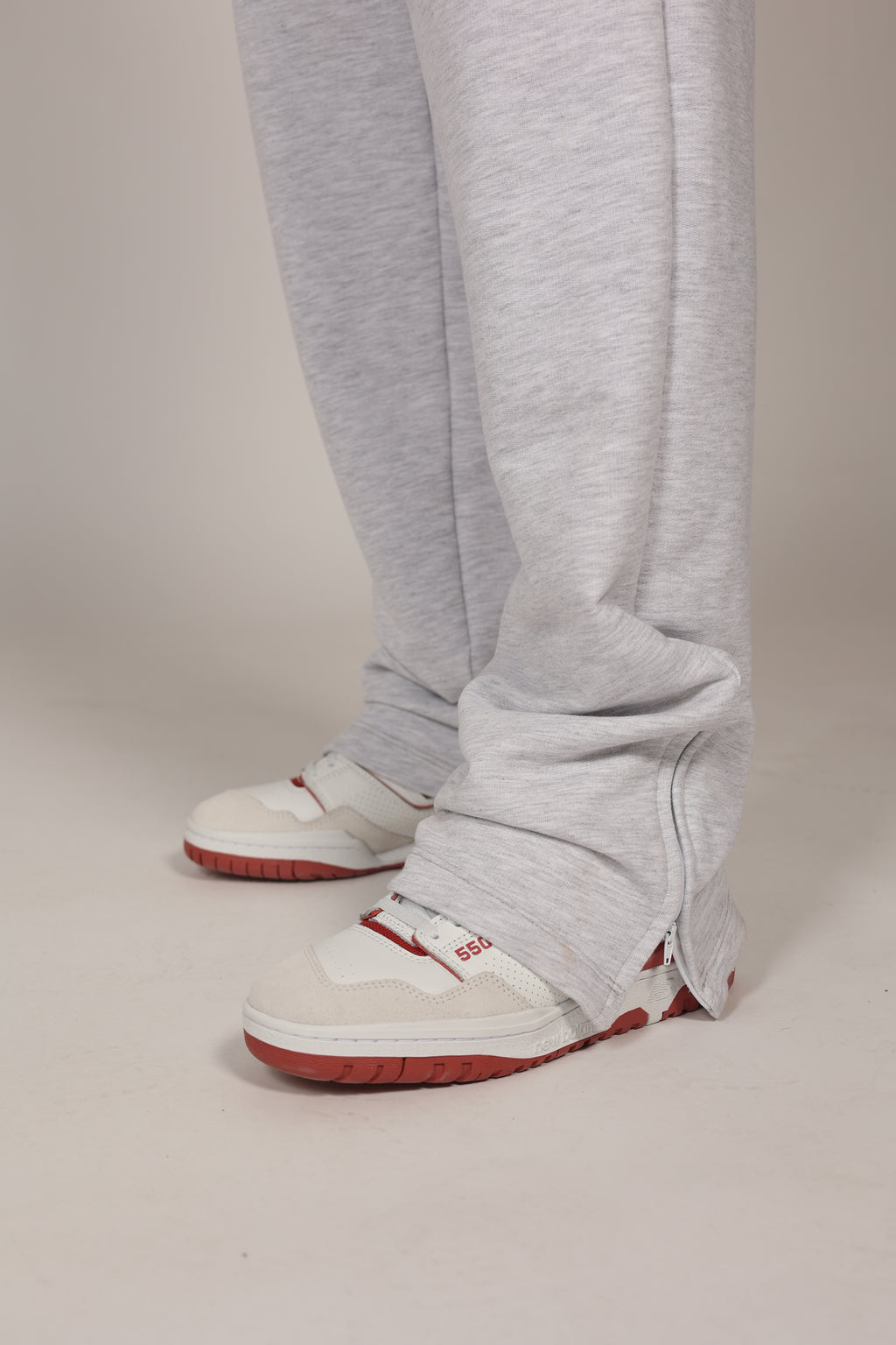 Hombre atlético vistiendo un pantalón deportivo jogger en color gris claro jaspeado. La prenda cuenta con una cintura elástica fruncida con cordón de ajuste. El corte es relajado y se muestra combinado con unos sneakers blancos con detalles rojos, destacando su caída moderna sobre el calzado en un fondo gris neutro o negro