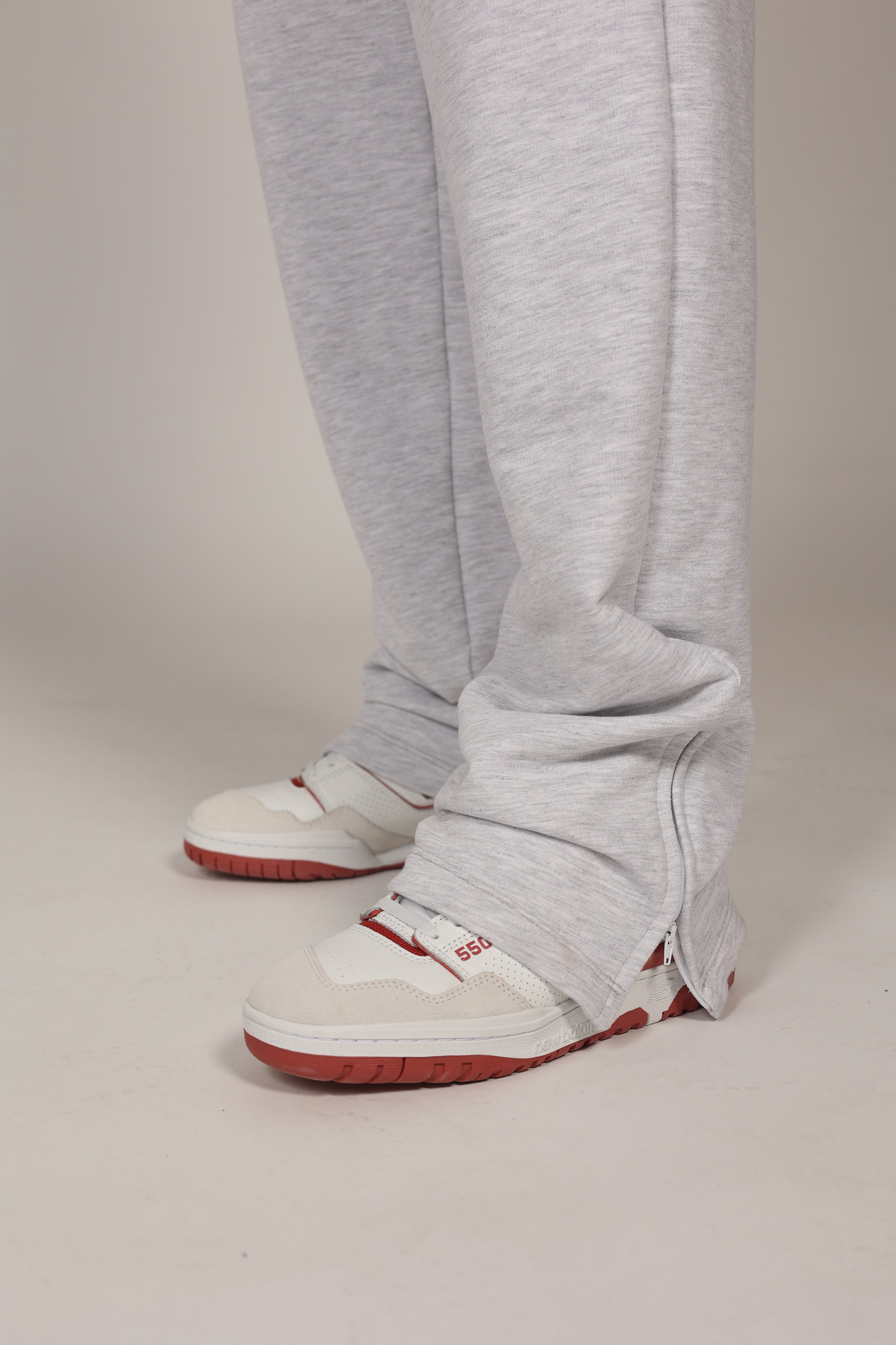 Hombre atlético vistiendo un pantalón deportivo jogger en color gris claro jaspeado. La prenda cuenta con una cintura elástica fruncida con cordón de ajuste. El corte es relajado y se muestra combinado con unos sneakers blancos con detalles rojos, destacando su caída moderna sobre el calzado en un fondo gris neutro o negro