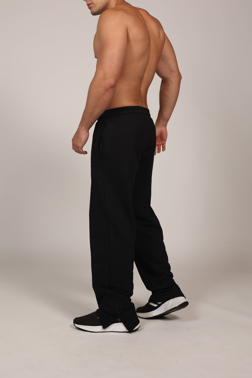Hombre atlético vistiendo un pantalón deportivo jogger en color gris claro jaspeado. La prenda cuenta con una cintura elástica fruncida con cordón de ajuste. El corte es relajado y se muestra combinado con unos sneakers blancos con detalles rojos, destacando su caída moderna sobre el calzado en un fondo gris neutro o negro