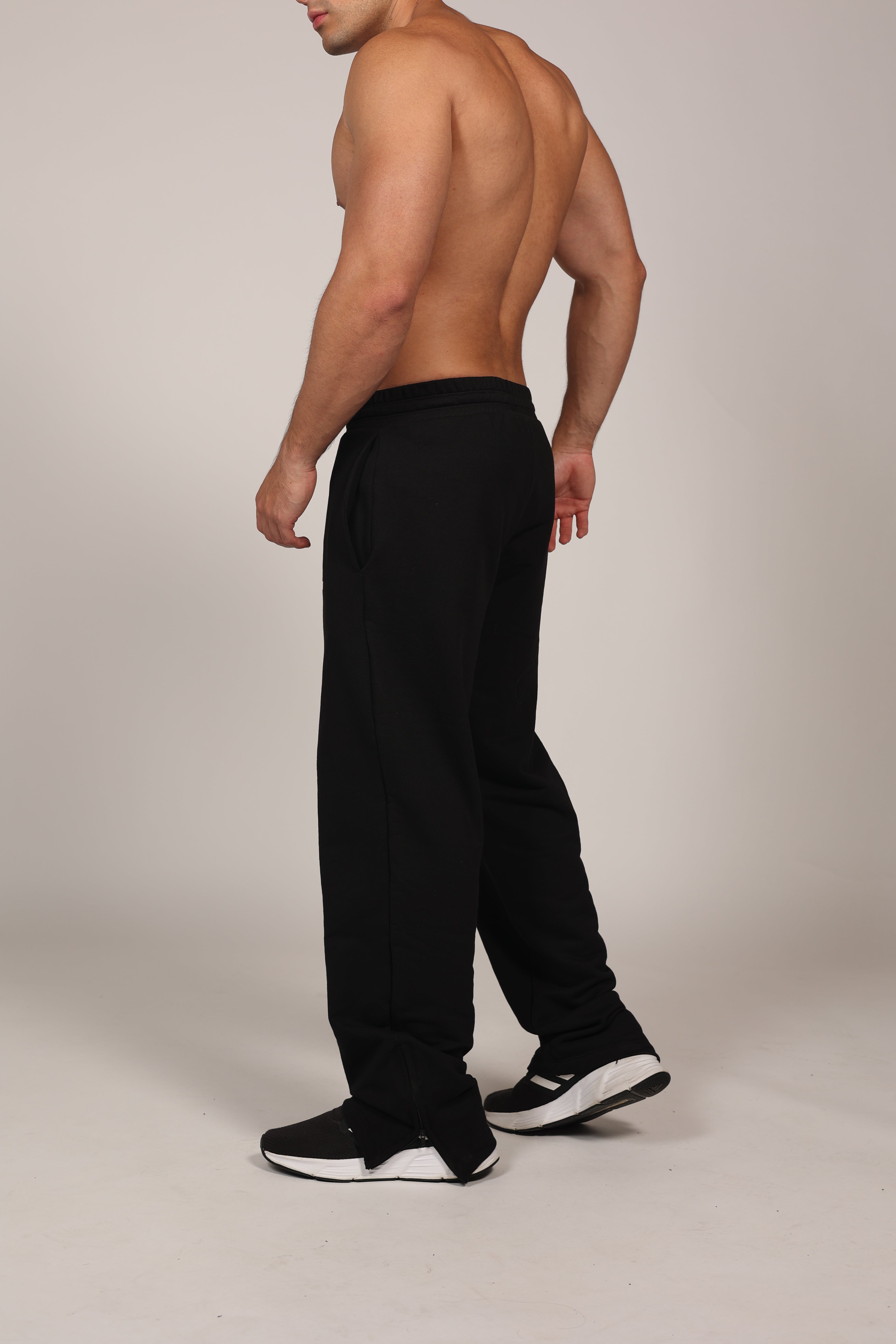 Hombre atlético vistiendo un pantalón deportivo jogger en color gris claro jaspeado. La prenda cuenta con una cintura elástica fruncida con cordón de ajuste. El corte es relajado y se muestra combinado con unos sneakers blancos con detalles rojos, destacando su caída moderna sobre el calzado en un fondo gris neutro o negro