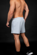 Modelo masculino atlético posando de perfil con pantaloneta deportiva color gris claro (azul hielo) de poliéster. El short tiene cintura elástica y cordón de ajuste negro. El modelo completa el outfit con medias y zapatillas de running negras, destacando un look moderno para gimnasio y atletismo sobre fondo oscuro.