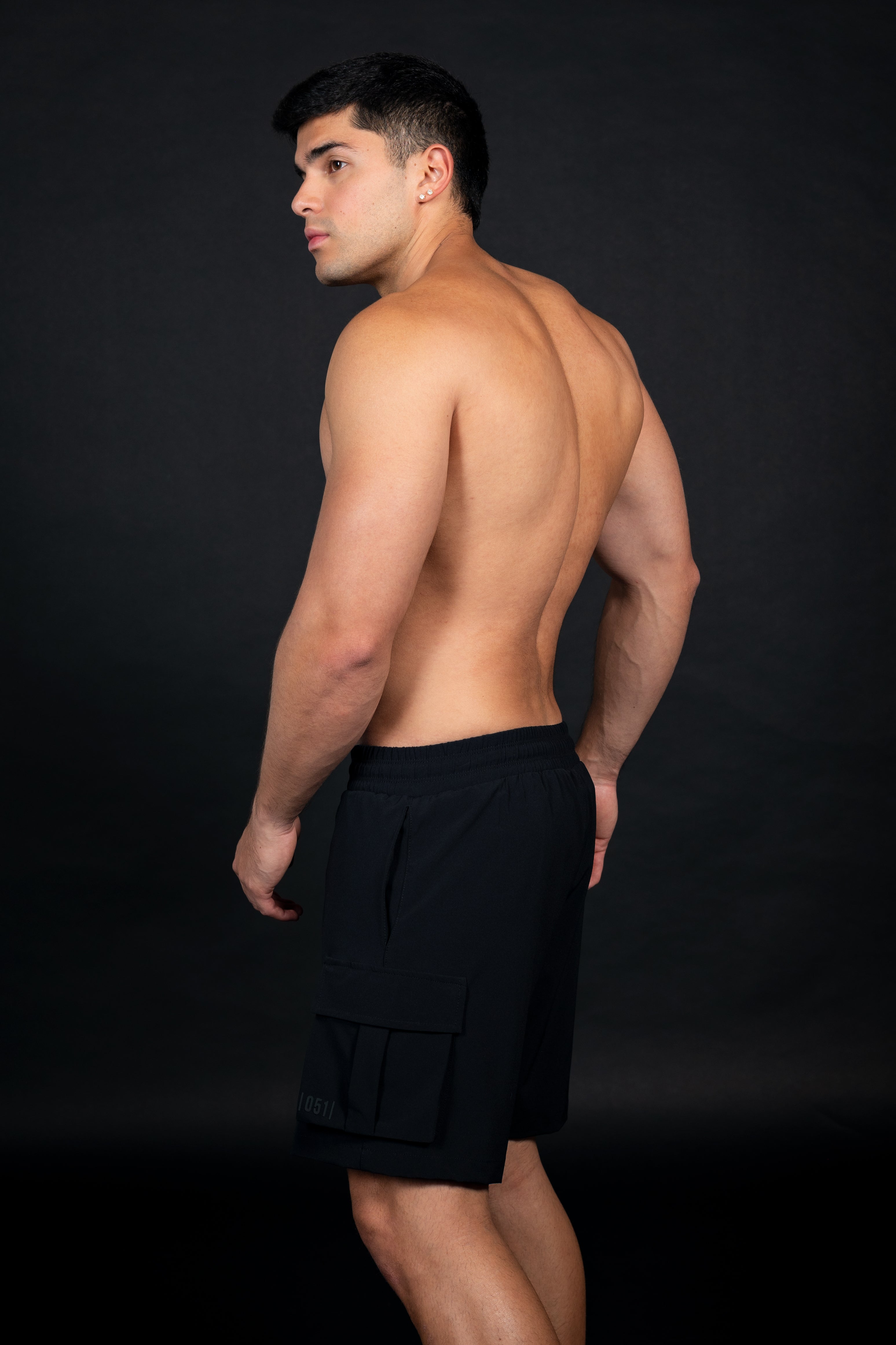Hombre atlético de perfil usando shorts tipo cargo de color negro, confeccionados en tela técnica ligera. La prenda cuenta con bolsillos laterales de gran tamaño, pretina elástica con cordón de ajuste marcado con letras blancas y una longitud por encima de la rodilla. El modelo posa sobre un fondo negro, destacando el corte moderno y funcional de la prenda.