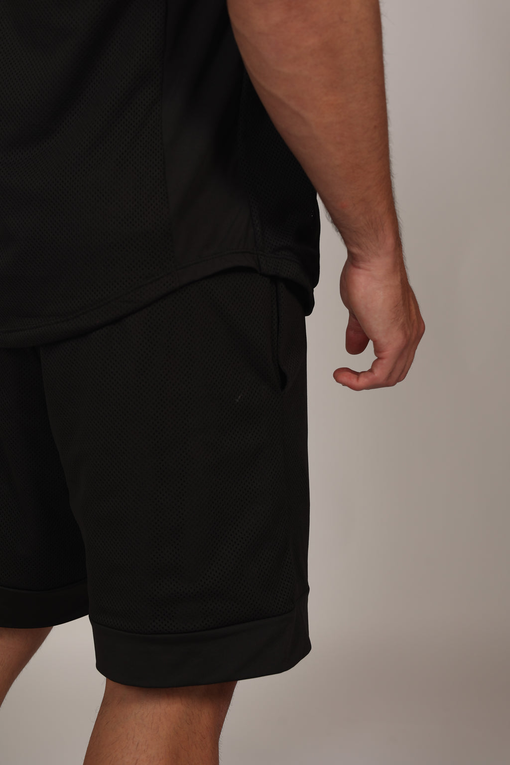 Primer plano de un short deportivo negro confeccionado en malla técnica con textura microperforada. Se aprecia la cintura elástica con cordón ajustable y un bolsillo lateral donde el modelo apoya su mano. En la parte inferior izquierda, destaca un parche cuadrado con branding sutil sobre el fondo de malla oscura.
