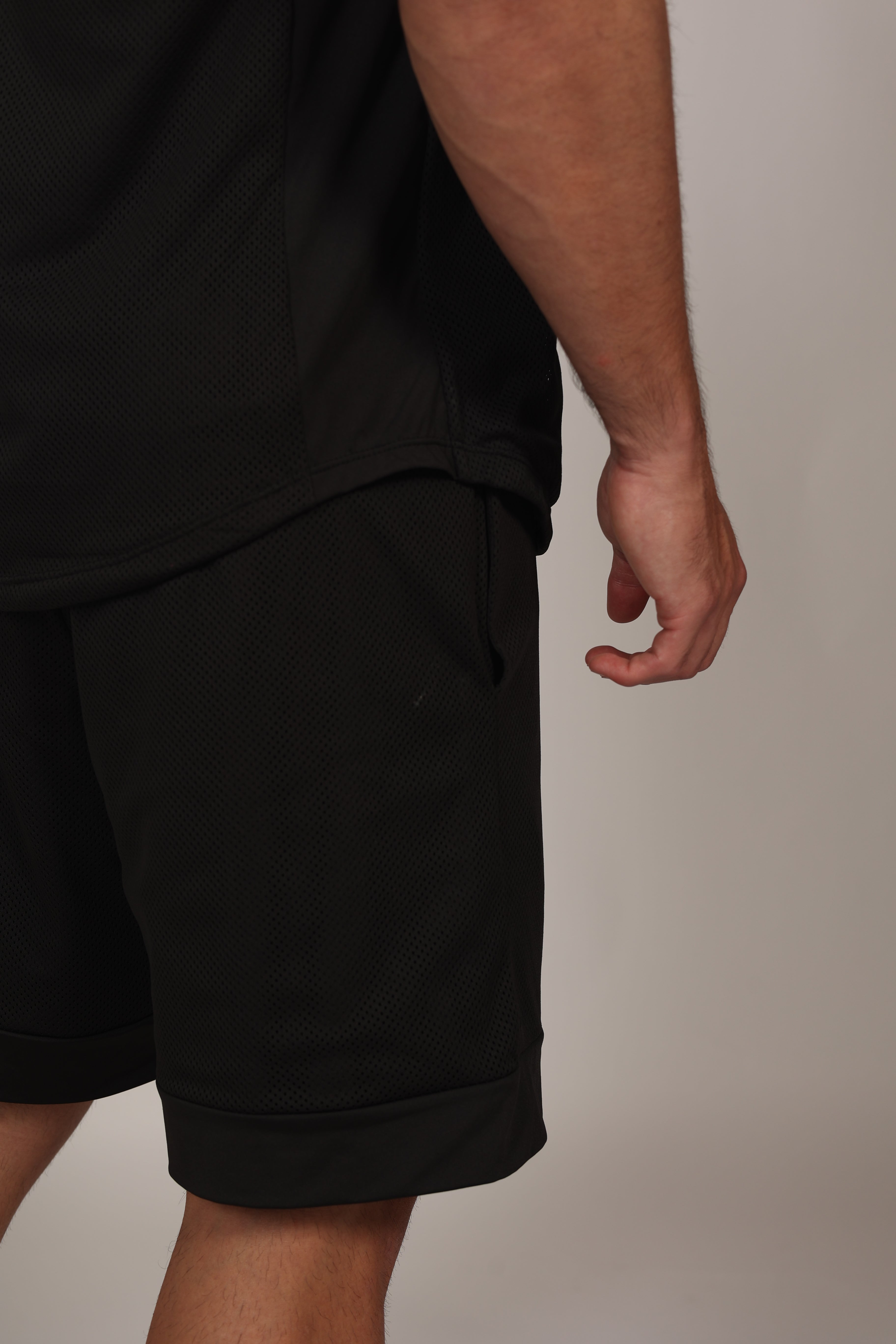 Primer plano de un short deportivo negro confeccionado en malla técnica con textura microperforada. Se aprecia la cintura elástica con cordón ajustable y un bolsillo lateral donde el modelo apoya su mano. En la parte inferior izquierda, destaca un parche cuadrado con branding sutil sobre el fondo de malla oscura.