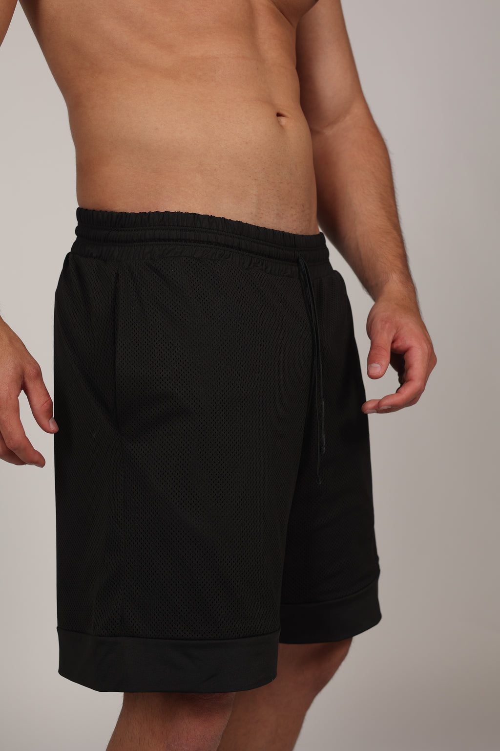 Primer plano de un short deportivo negro confeccionado en malla técnica con textura microperforada. Se aprecia la cintura elástica con cordón ajustable y un bolsillo lateral donde el modelo apoya su mano. En la parte inferior izquierda, destaca un parche cuadrado con branding sutil sobre el fondo de malla oscura.