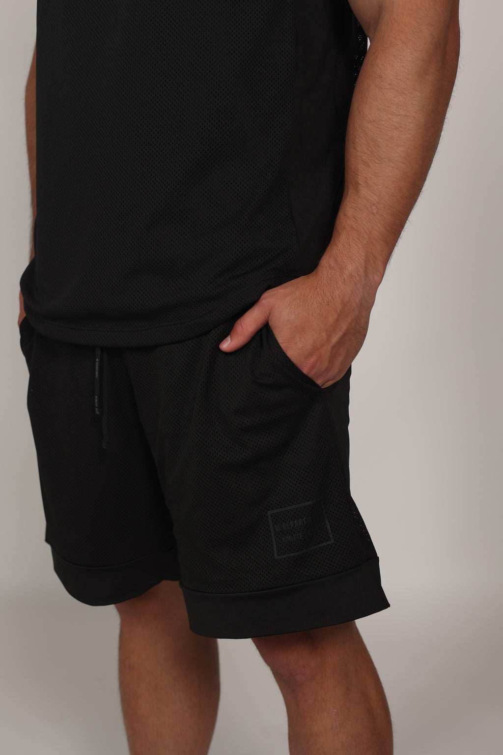 Primer plano de un short deportivo negro confeccionado en malla técnica con textura microperforada. Se aprecia la cintura elástica con cordón ajustable y un bolsillo lateral donde el modelo apoya su mano. En la parte inferior izquierda, destaca un parche cuadrado con branding sutil sobre el fondo de malla oscura.