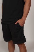 Primer plano de un short deportivo negro confeccionado en malla técnica con textura microperforada. Se aprecia la cintura elástica con cordón ajustable y un bolsillo lateral donde el modelo apoya su mano. En la parte inferior izquierda, destaca un parche cuadrado con branding sutil sobre el fondo de malla oscura.