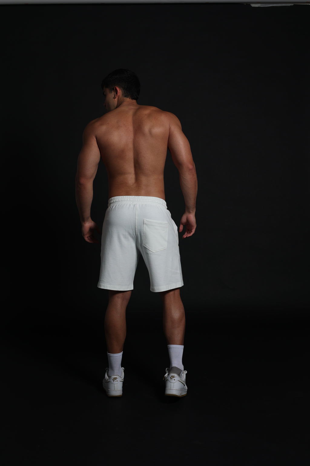 Hombre atlético de perfil vistiendo shorts de algodón elástico en color ivory o negro . La imagen destaca el ajuste cómodo por encima de la rodilla, la cintura elástica fruncida con cordón negro y el diseño de bolsillos funcionales con cremallera, ideal para un estilo de vida deportivo y urbano sobre un fondo negro