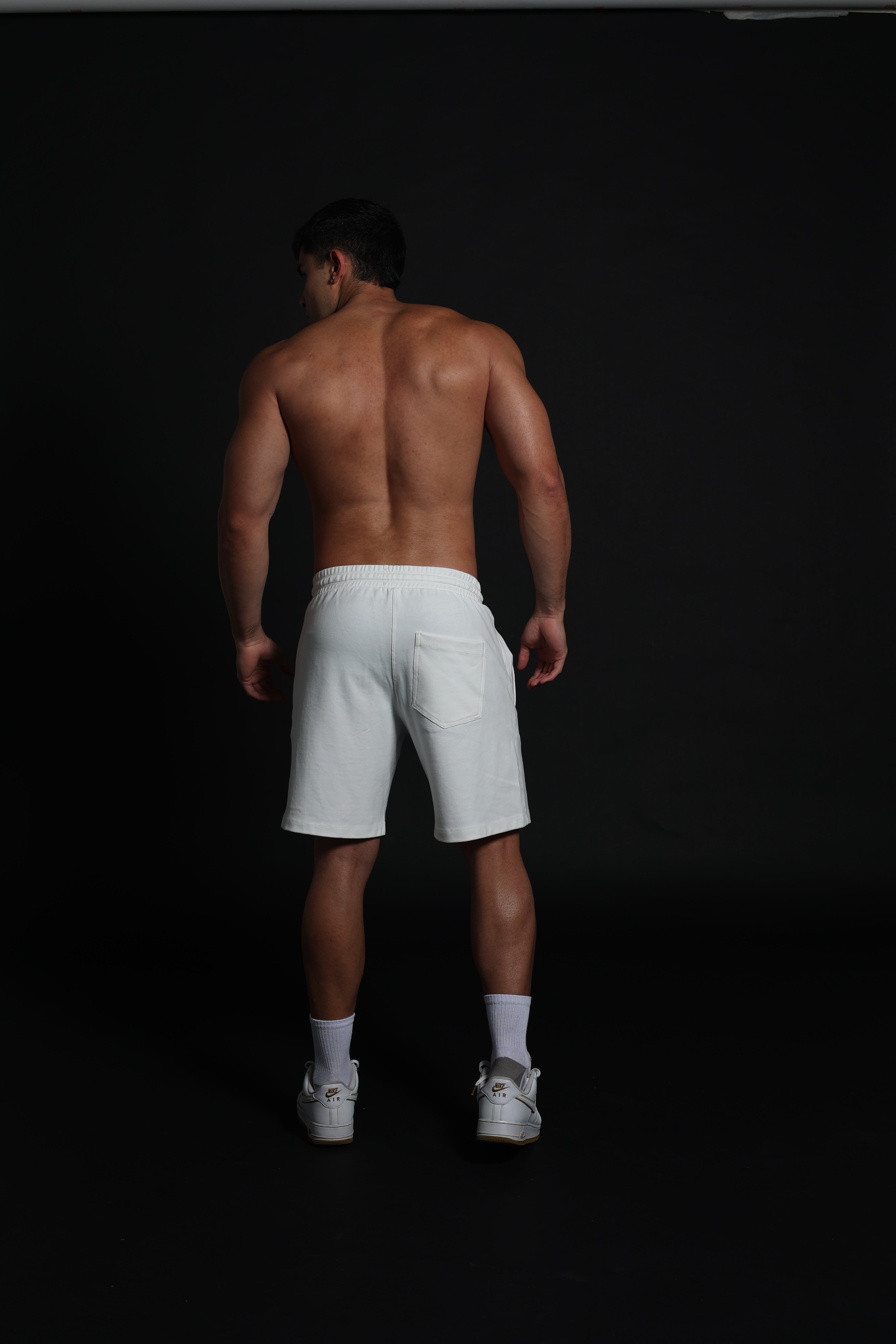 Hombre atlético de perfil vistiendo shorts de algodón elástico en color ivory o negro . La imagen destaca el ajuste cómodo por encima de la rodilla, la cintura elástica fruncida con cordón negro y el diseño de bolsillos funcionales con cremallera, ideal para un estilo de vida deportivo y urbano sobre un fondo negro