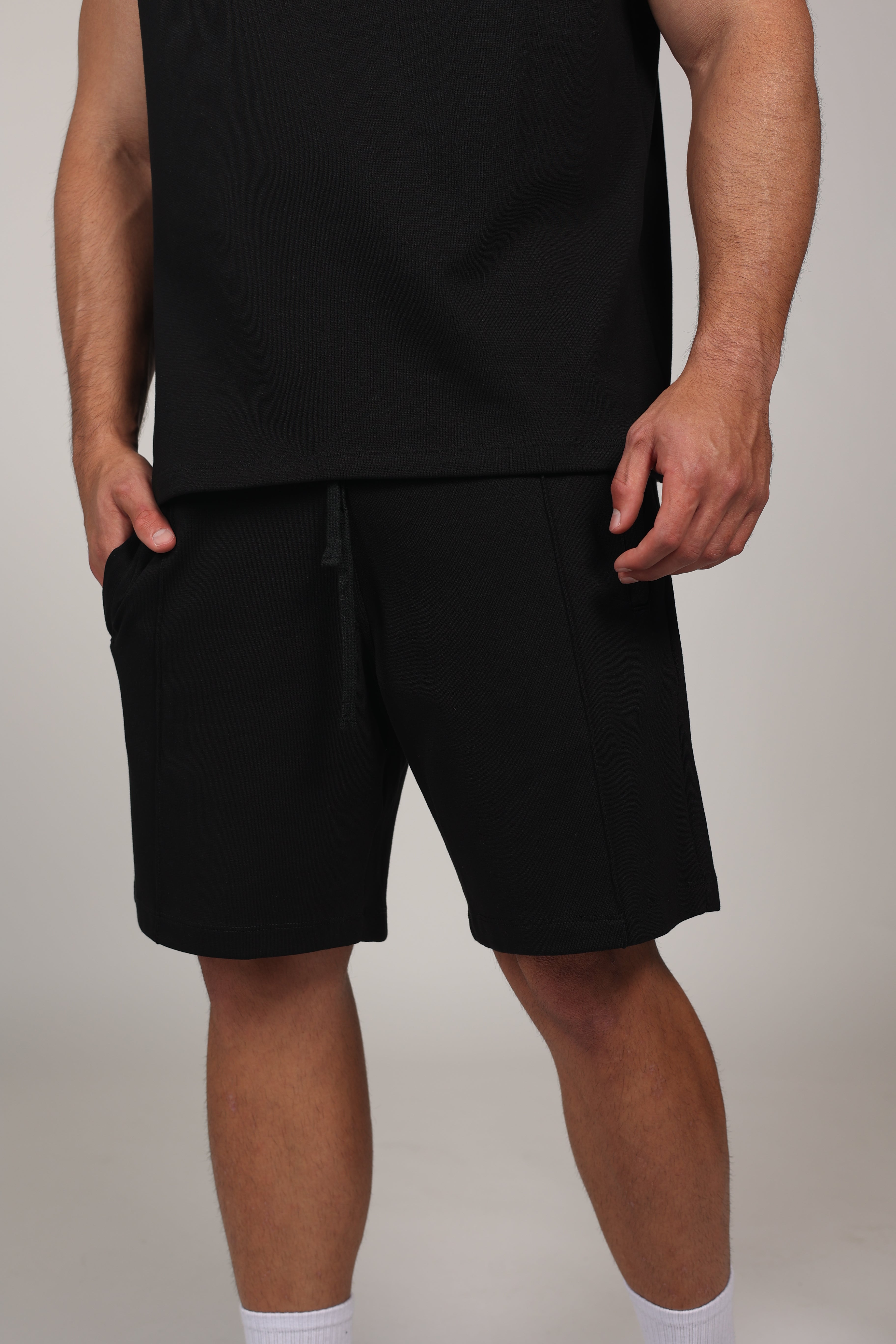 Hombre atlético de perfil vistiendo shorts de algodón elástico en color ivory o negro . La imagen destaca el ajuste cómodo por encima de la rodilla, la cintura elástica fruncida con cordón negro y el diseño de bolsillos funcionales con cremallera, ideal para un estilo de vida deportivo y urbano sobre un fondo negro