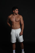Hombre atlético de perfil vistiendo shorts de algodón elástico en color ivory o negro . La imagen destaca el ajuste cómodo por encima de la rodilla, la cintura elástica fruncida con cordón negro y el diseño de bolsillos funcionales con cremallera, ideal para un estilo de vida deportivo y urbano sobre un fondo negro