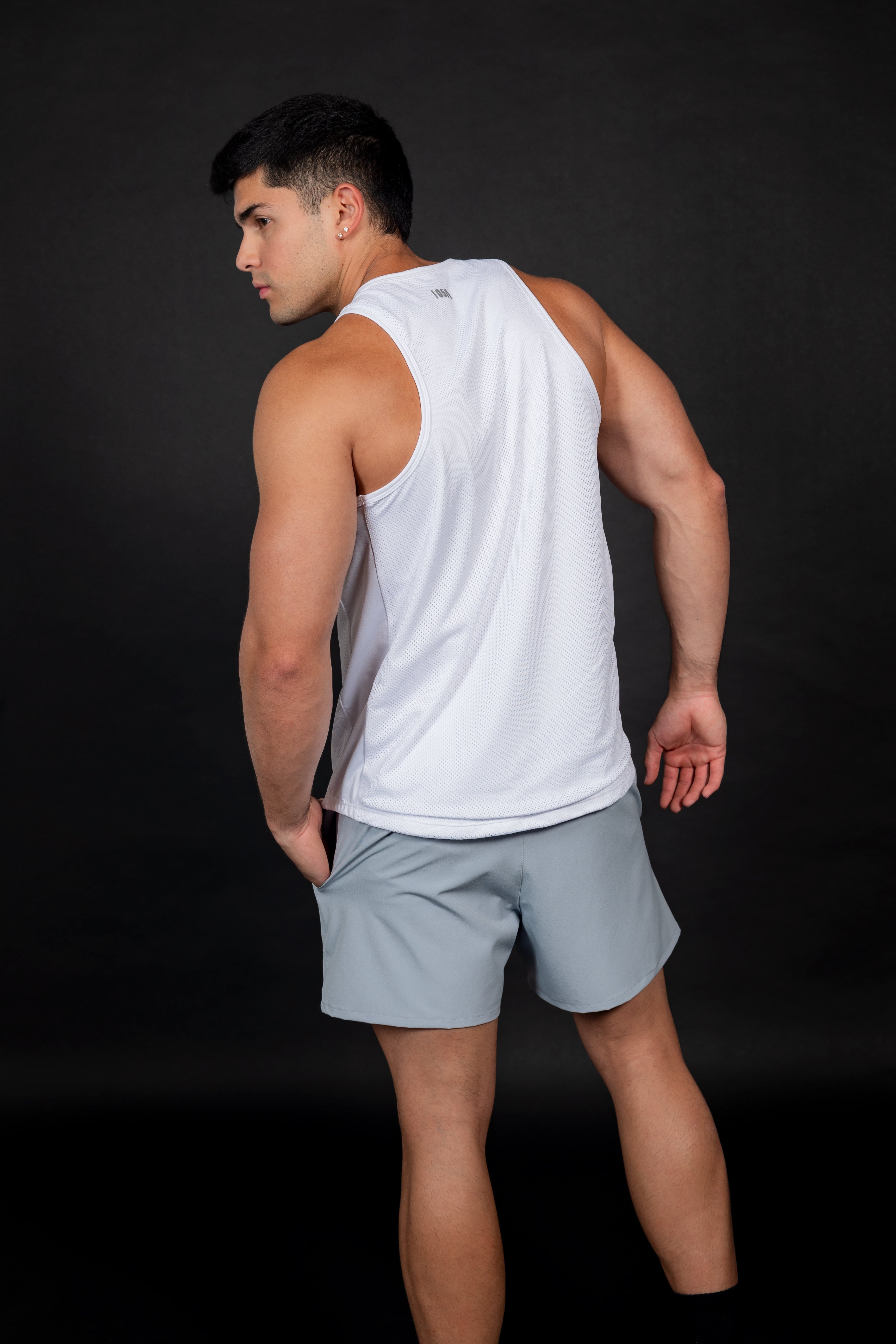 Hombre atlético de frente vistiendo un tank top blanco de malla técnica con textura microperforada. La prenda tiene un cuello redondo y sisas amplias, combinada con shorts gris claro. El diseño resalta la musculatura de los brazos y hombros sobre un fondo negro, evocando una estética de jugador de basketball profesiona